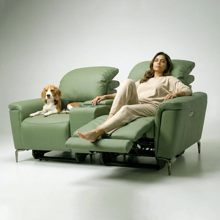 A woman in beige loungewear reclines on a green upholstered recliner chair, with a brown and white dog resting on the adjacent matching chair, set against a clean light gray studio background.