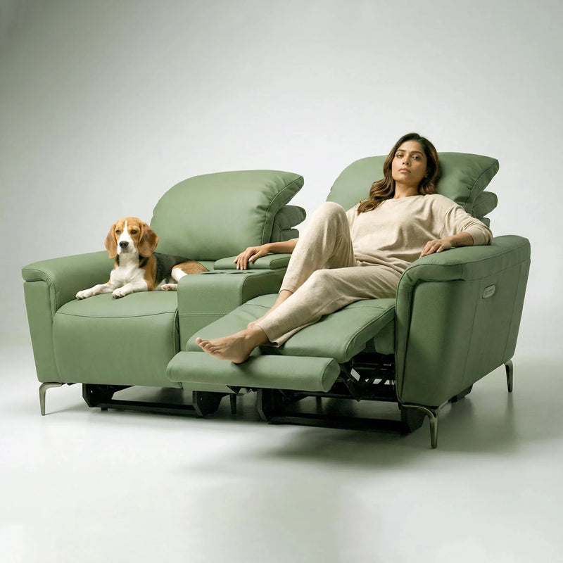 A woman in beige loungewear reclines on a green upholstered recliner chair, with a brown and white dog resting on the adjacent matching chair, set against a clean light gray studio background.